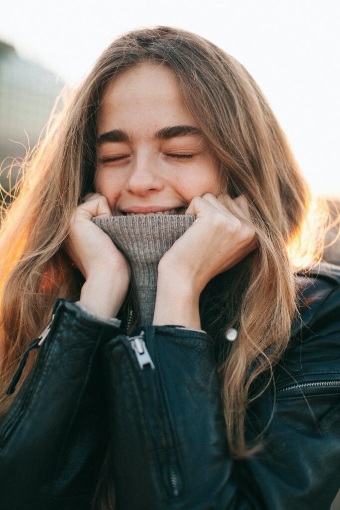 Happy young female with long hair in warm turtleneck and leather jacket enjoying sunny day with closed eyes on street